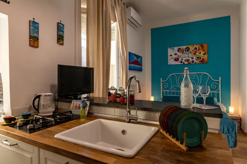 La cucina: il lavello, i fornelli a gas e i piatti colorati sulla penisola; dietro, il letto matrimoniale sulla parete turchese.